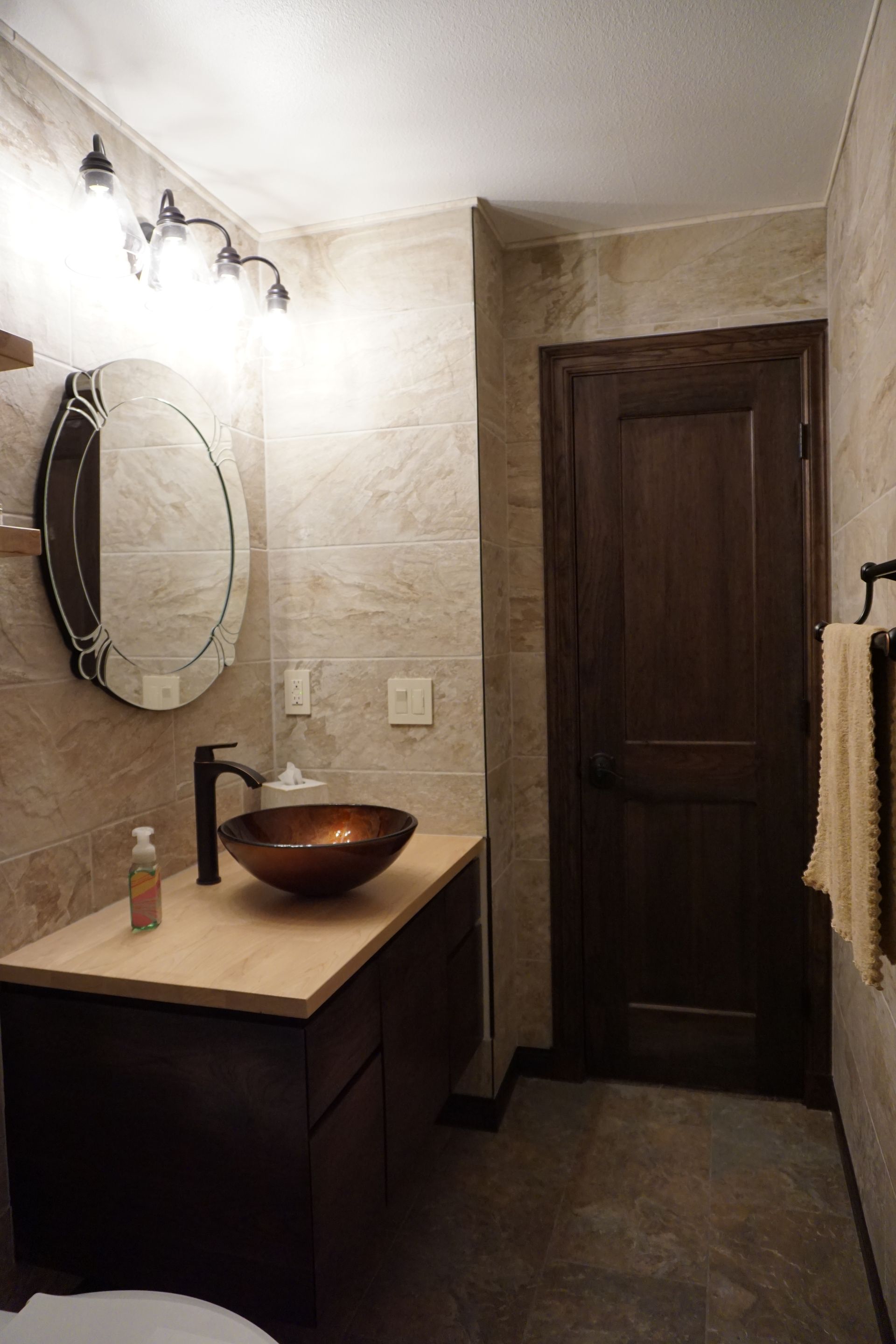 Bathroom with brown vanity, copper sink, oval mirror, dark wooden door, and stone tile walls.