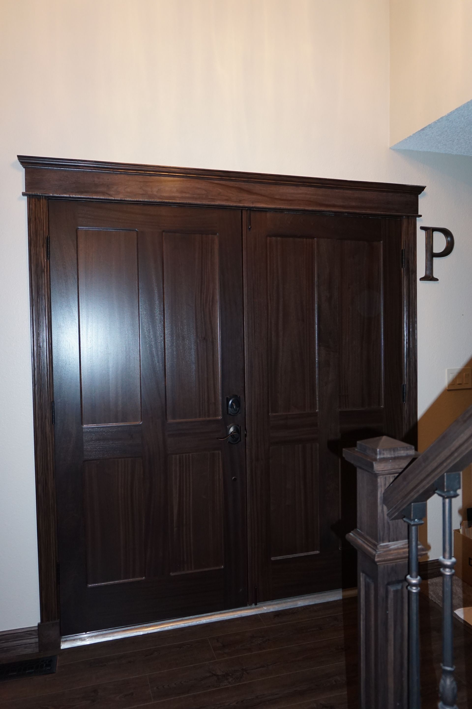 Double dark wooden doors with trim and a handle. The doors are next to a staircase.