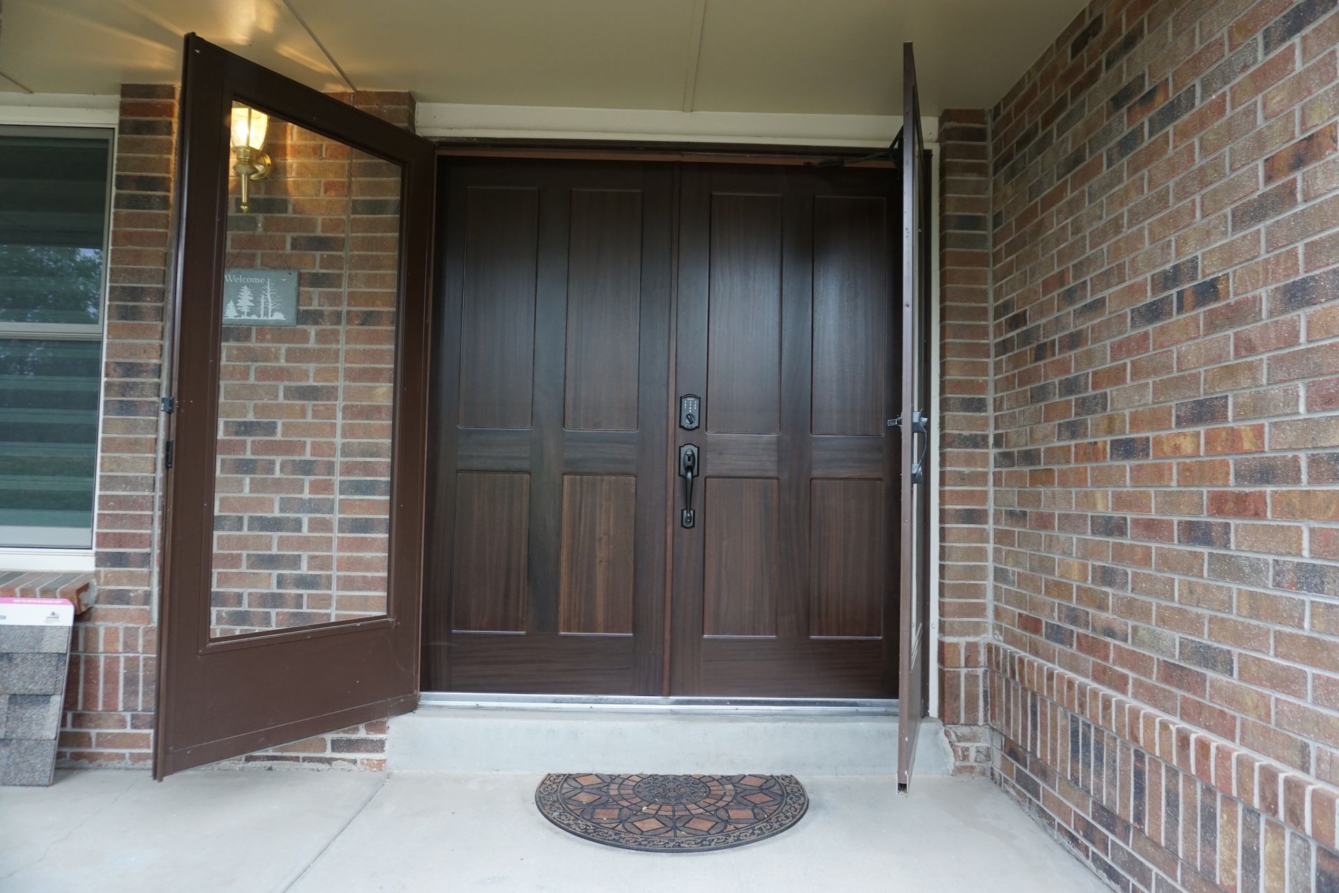Brown double doors open on a brick building with a welcome mat in front.