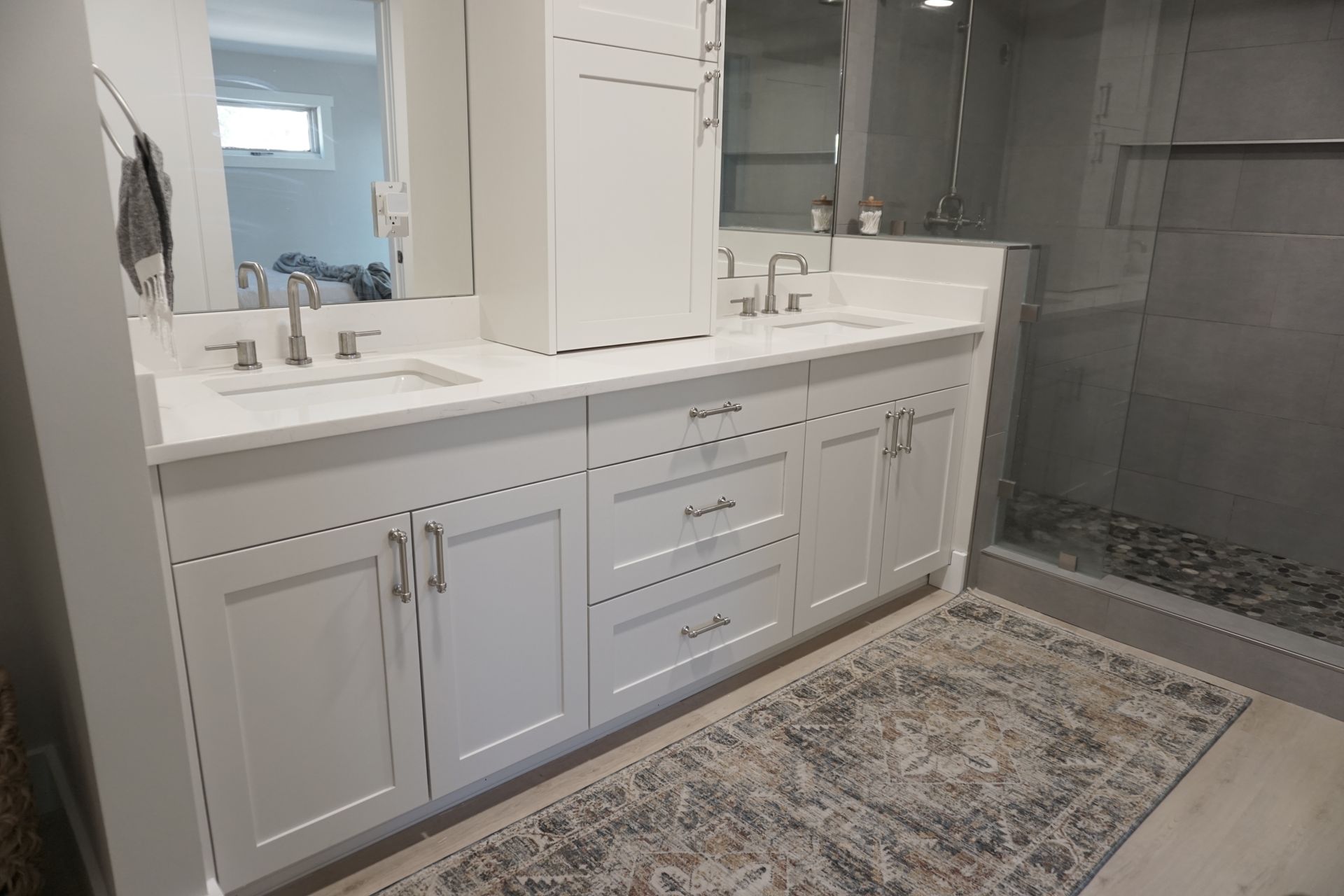 White bathroom vanity with dual sinks, cabinets, and a rug. Shower with glass door.