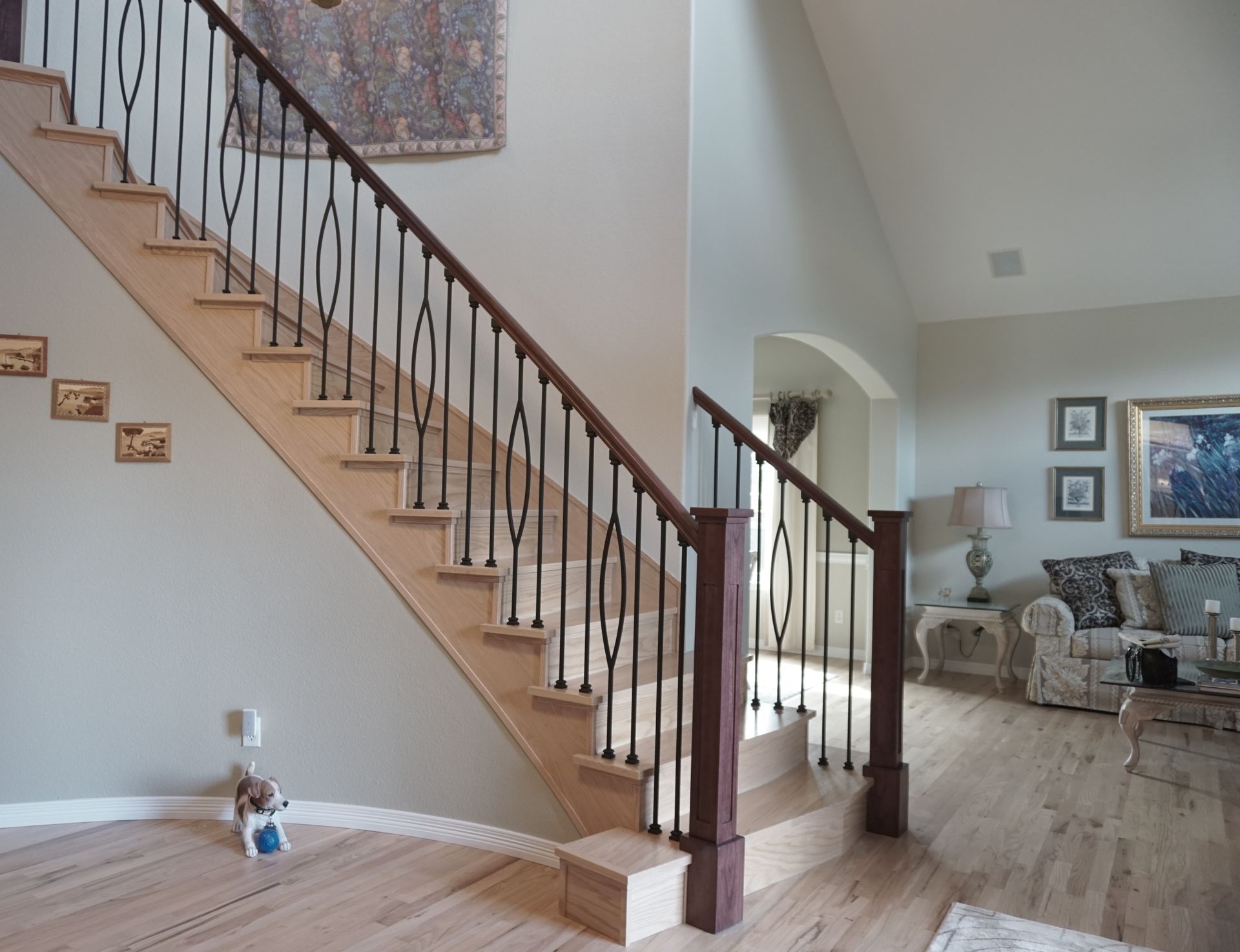Staircase with light wood steps and dark iron railing. Beige walls, hardwood floors, and a living room in the background.