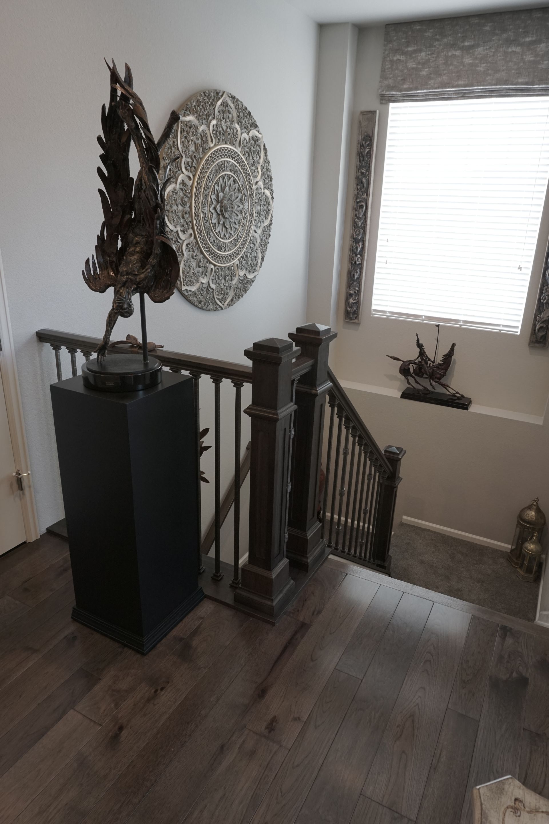 Stairwell with dark wood floors and banister. Sculpture on black pedestal, decorative art on wall, window with blinds.