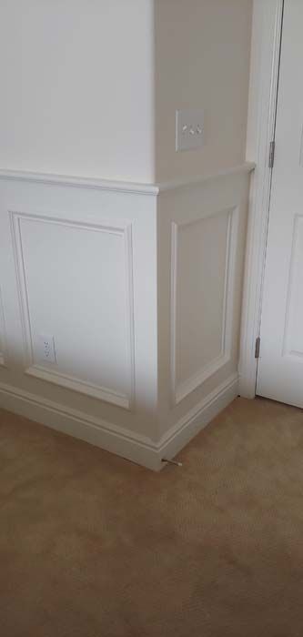 White paneling on corner wall and baseboard, with electrical outlet. Beige carpet. Door on right.
