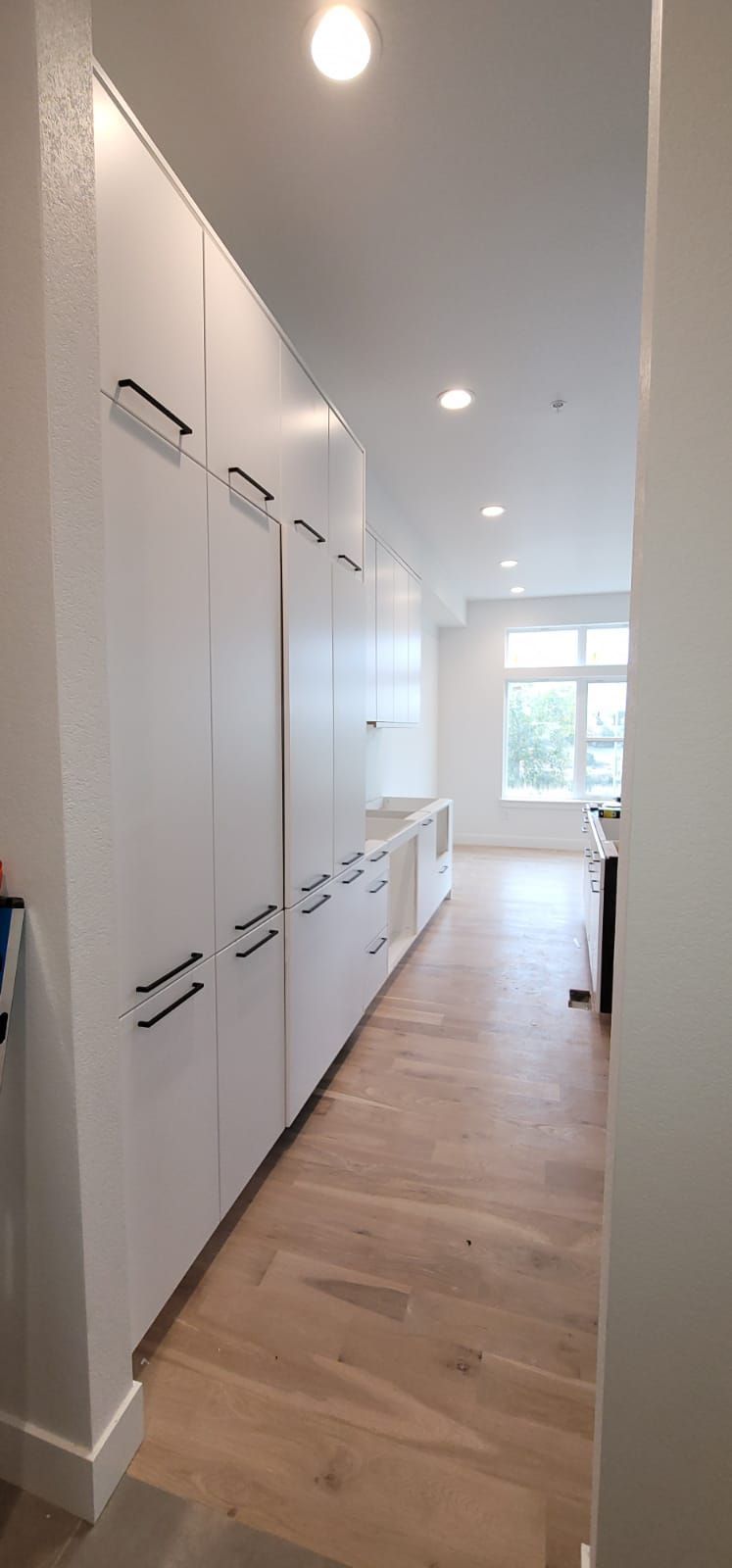 A long, narrow kitchen with white cabinets, light wood floors, and a bright window at the end.