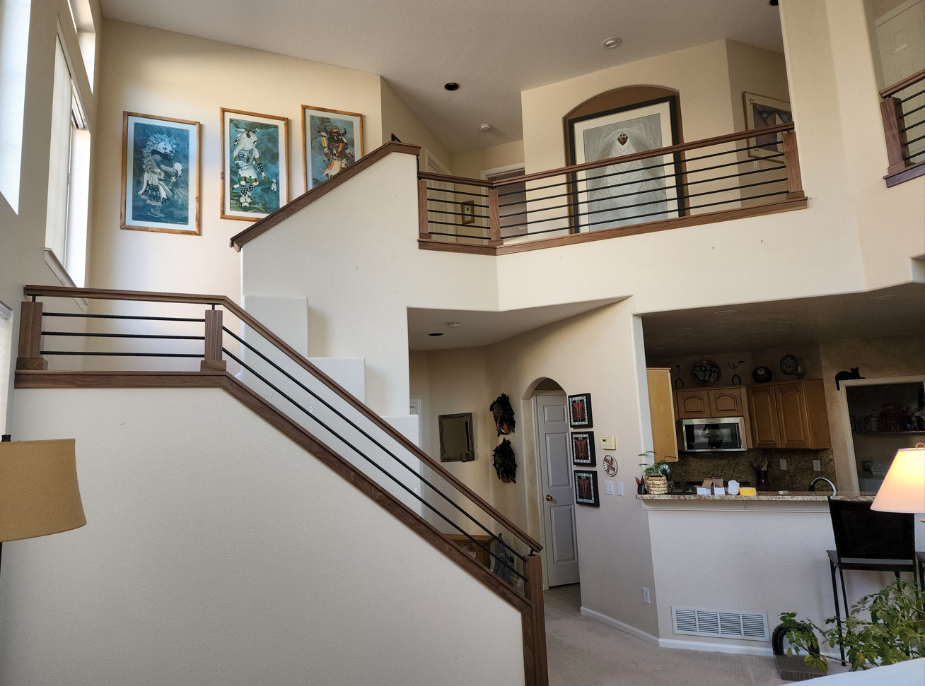 Two-story home interior with staircase and balcony. White walls, brown railings, artwork, and kitchen visible in the background.