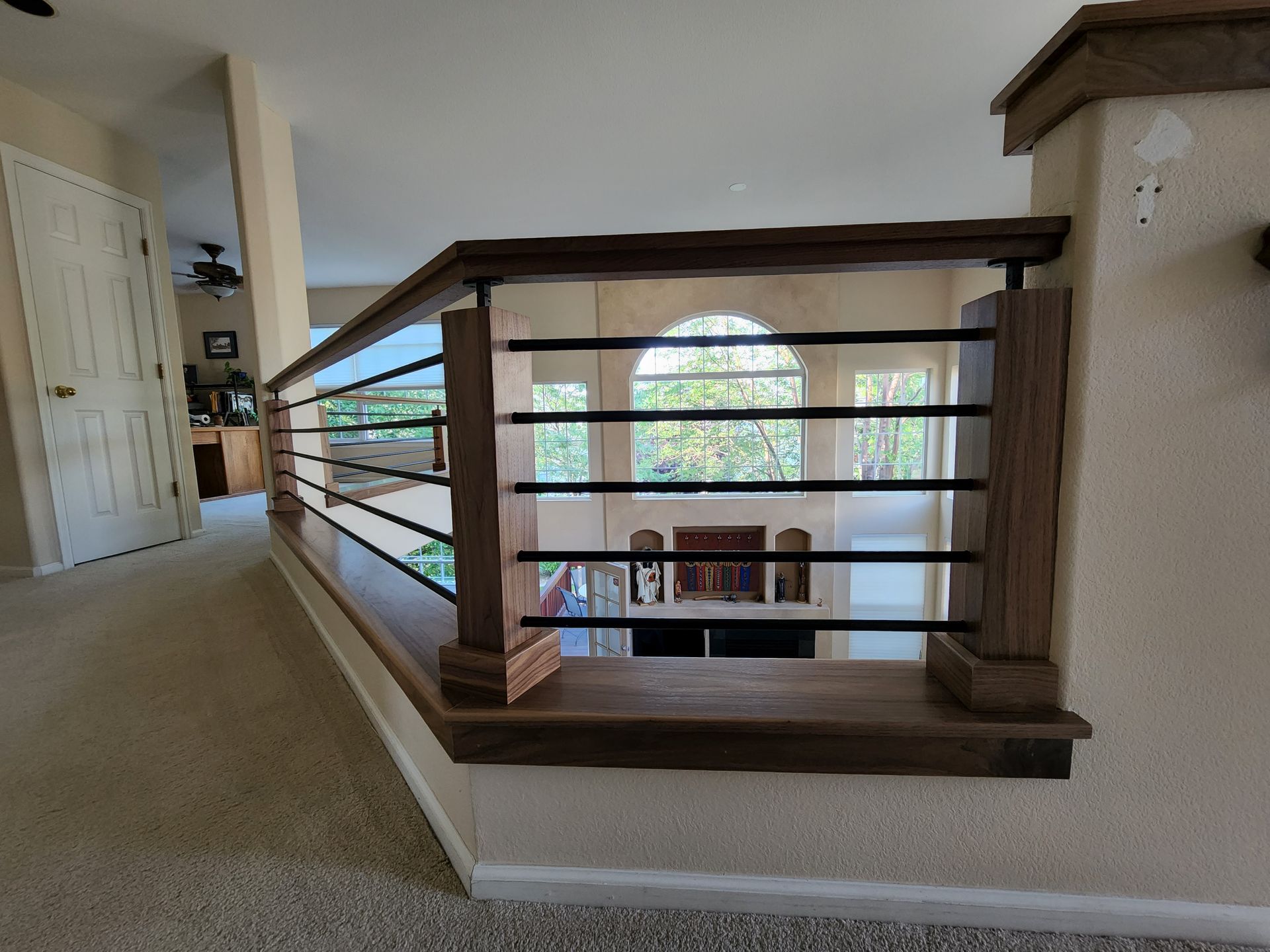 Interior view: wooden railing with black bars overlooks a living room with arched windows.
