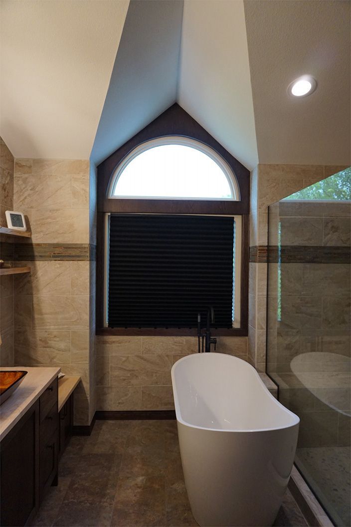 Bathroom with freestanding tub under an arched window with dark blinds. Beige tiled walls, brown trim.