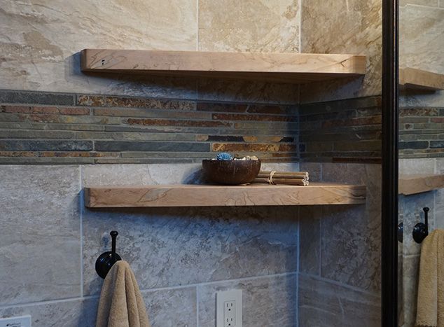 Two wooden shelves in a tiled bathroom corner holding a bowl and rolled towels; towels hang on hooks below.
