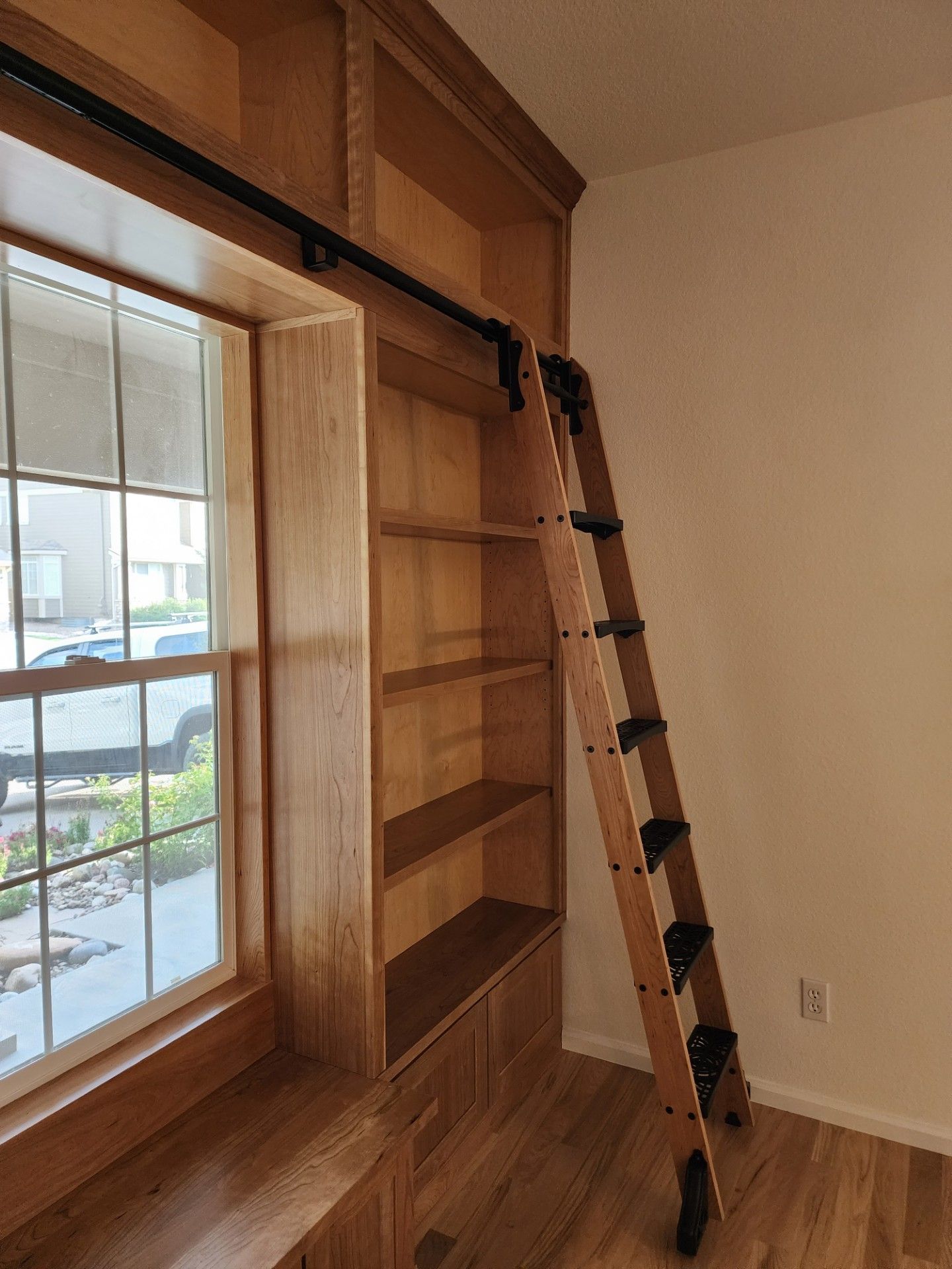 Wooden bookcase with rolling ladder beside a window with a bench.