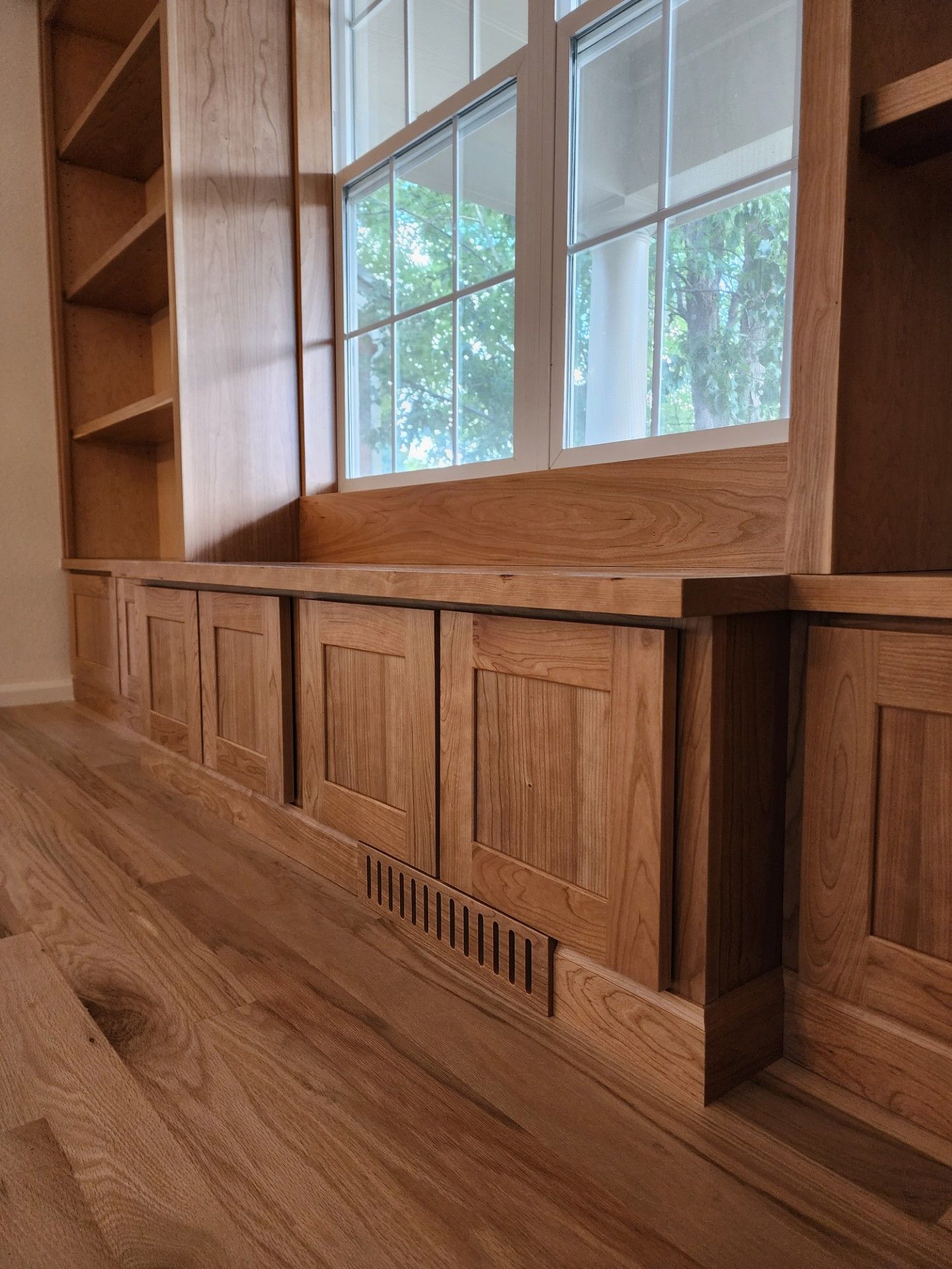 Wooden built-in storage and window seat with cabinets and bookshelves. Hardwood floor. Window with trees visible.