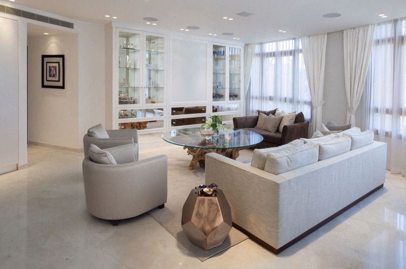 Elegant white living room with light-colored furniture, glass-topped table, glass display cabinets, and sheer white curtains.