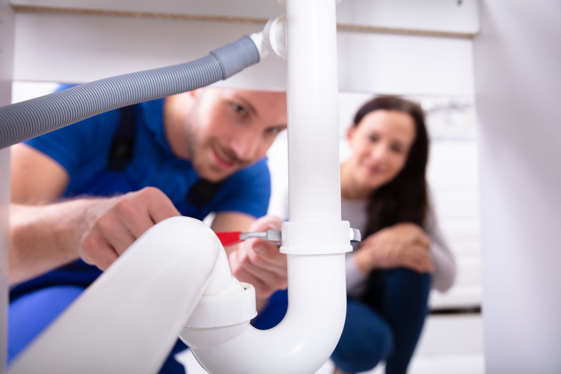 A plumber is fixing a sink pipe while a woman sits in the background.