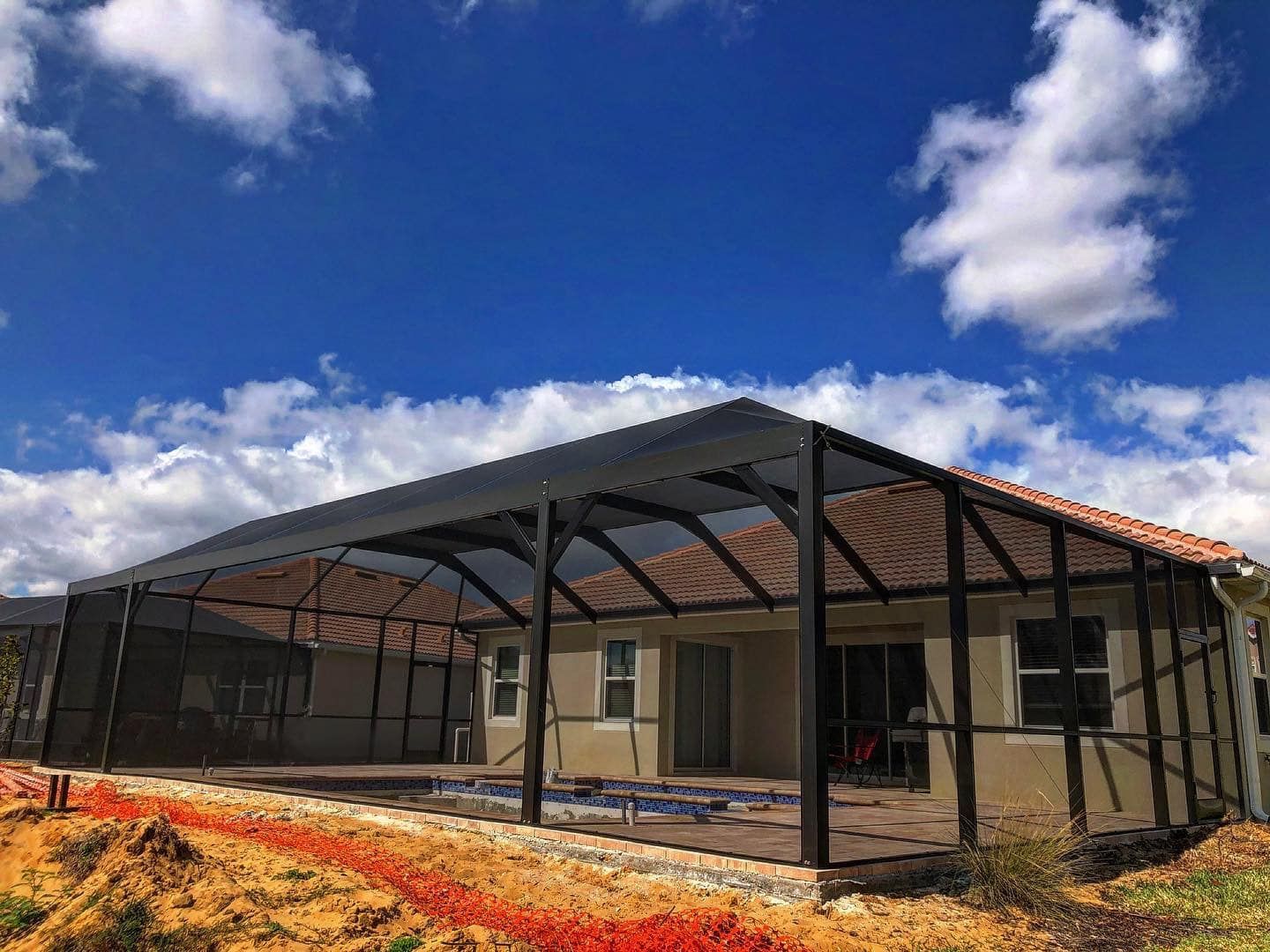 Screened-in patio enclosure over a pool. Black frame with tan house and blue sky.