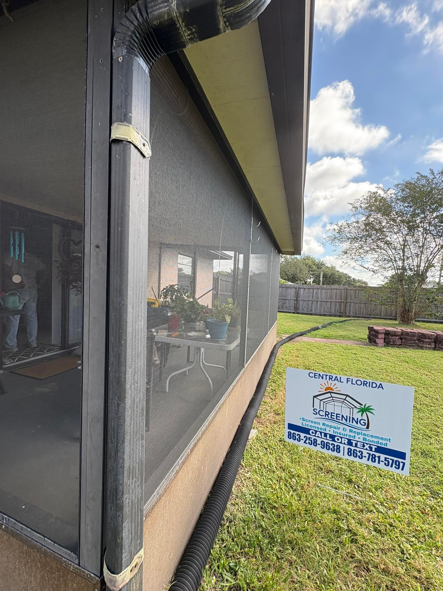 Screened-in porch with black frame, tan trim, and a gutter, viewed from a grassy yard with a sign.