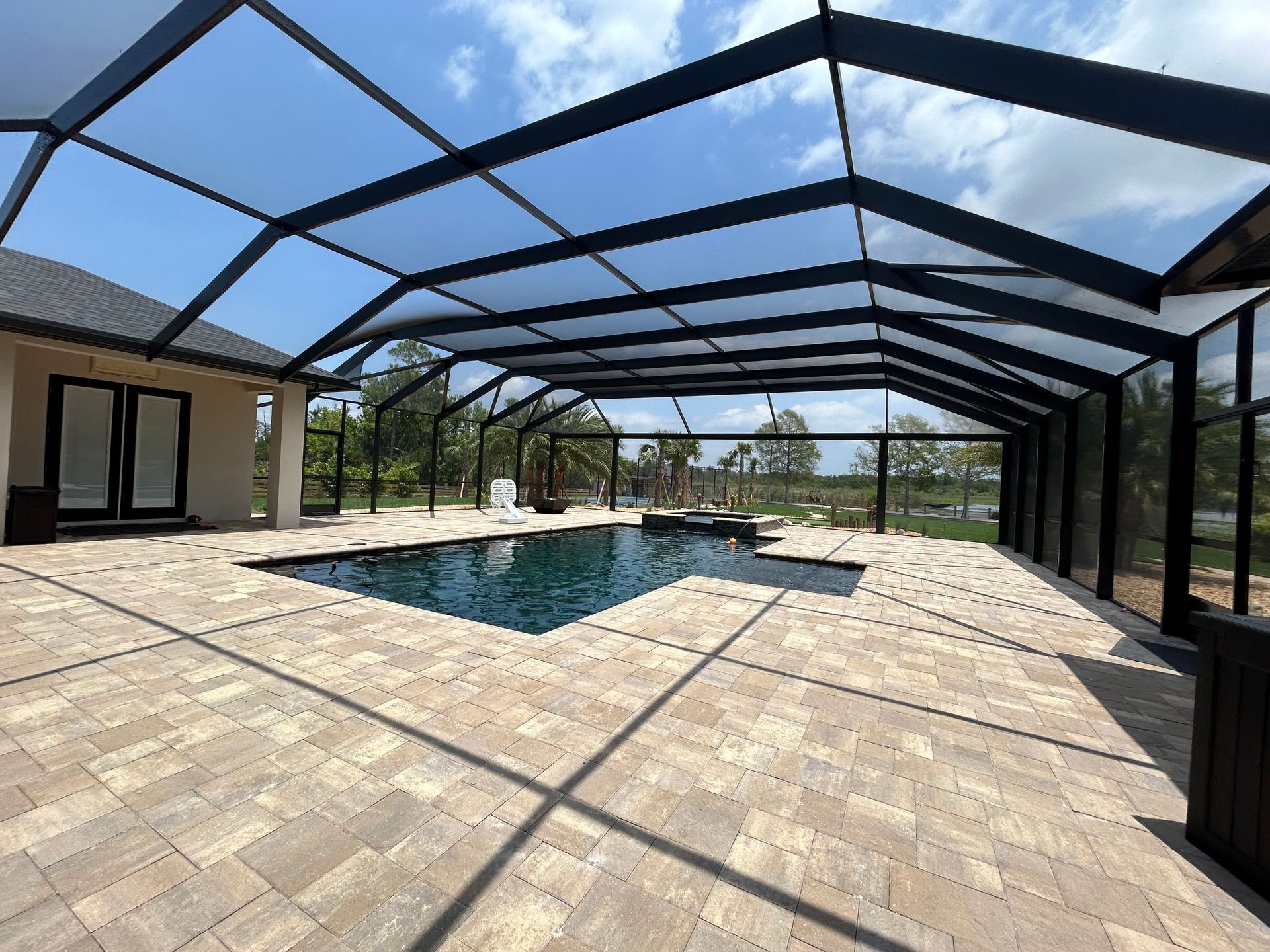 Pool area with a screened enclosure. Paver deck surrounds a dark pool; blue sky visible above.