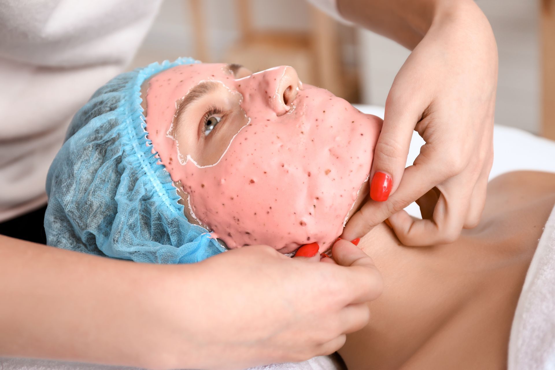 Woman receiving facial mask treatment in a spa, wearing blue hair cap.