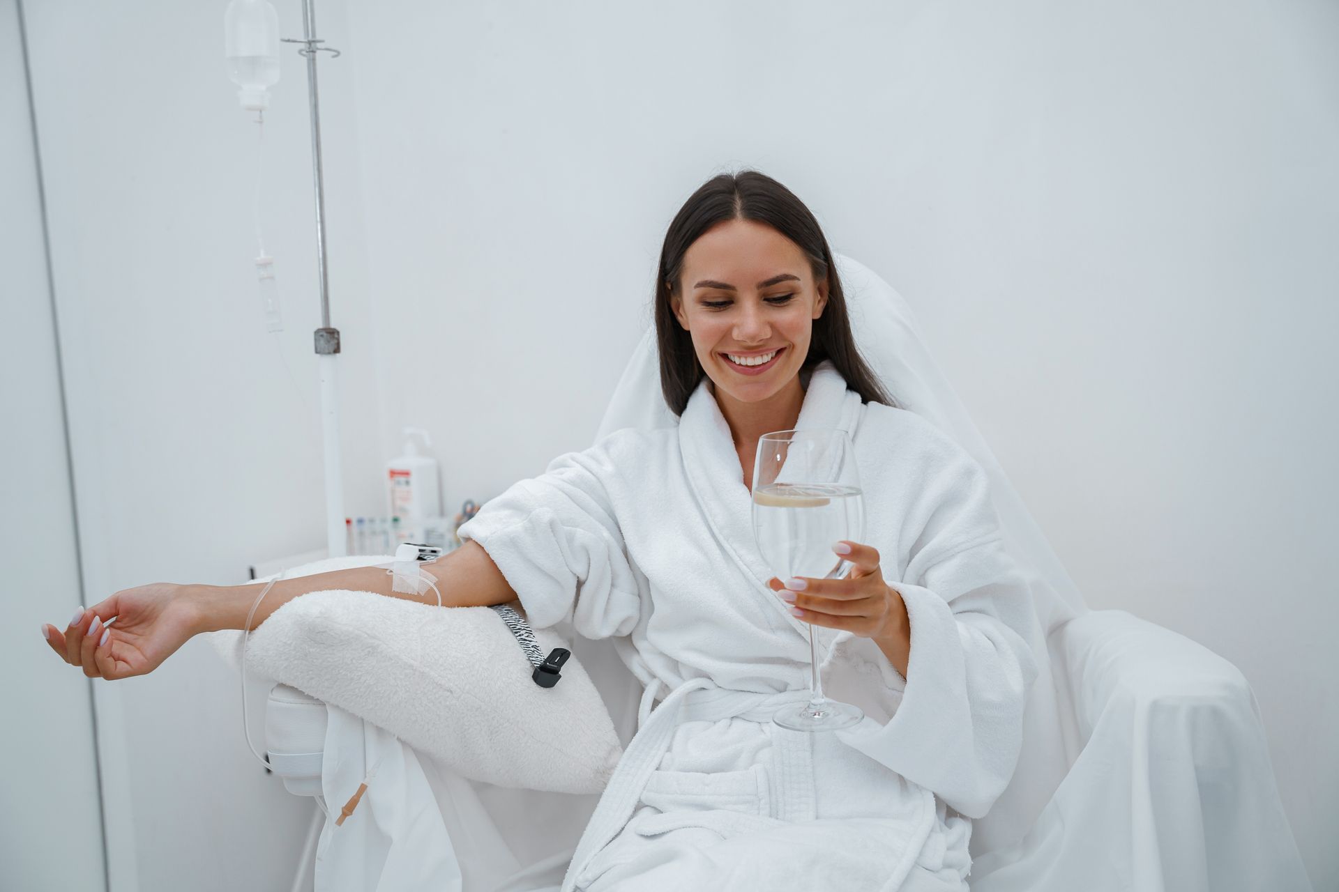 Woman in a white robe sitting on a bed, smiling and holding a glass in a bright room