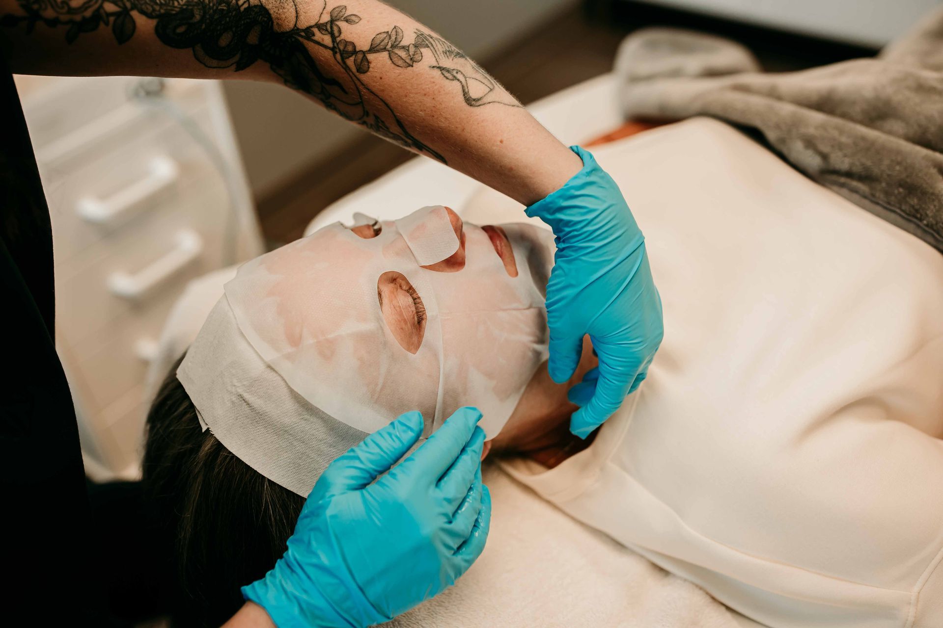 Person receiving a facial mask application. Hands in blue gloves place mask on face. White towels on surface.
