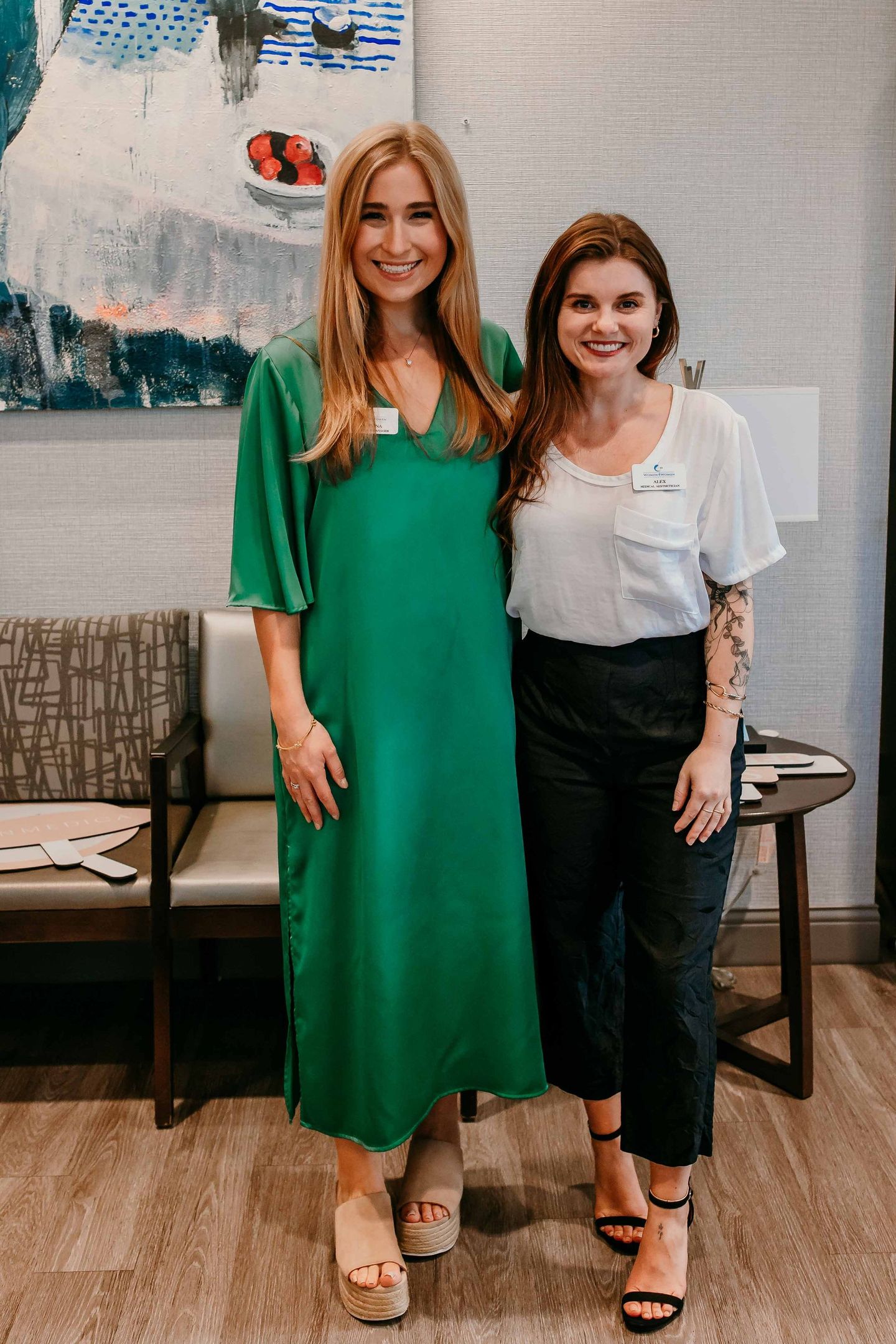 Two women are posing for a picture in a waiting room . one of the women is wearing a green dress.