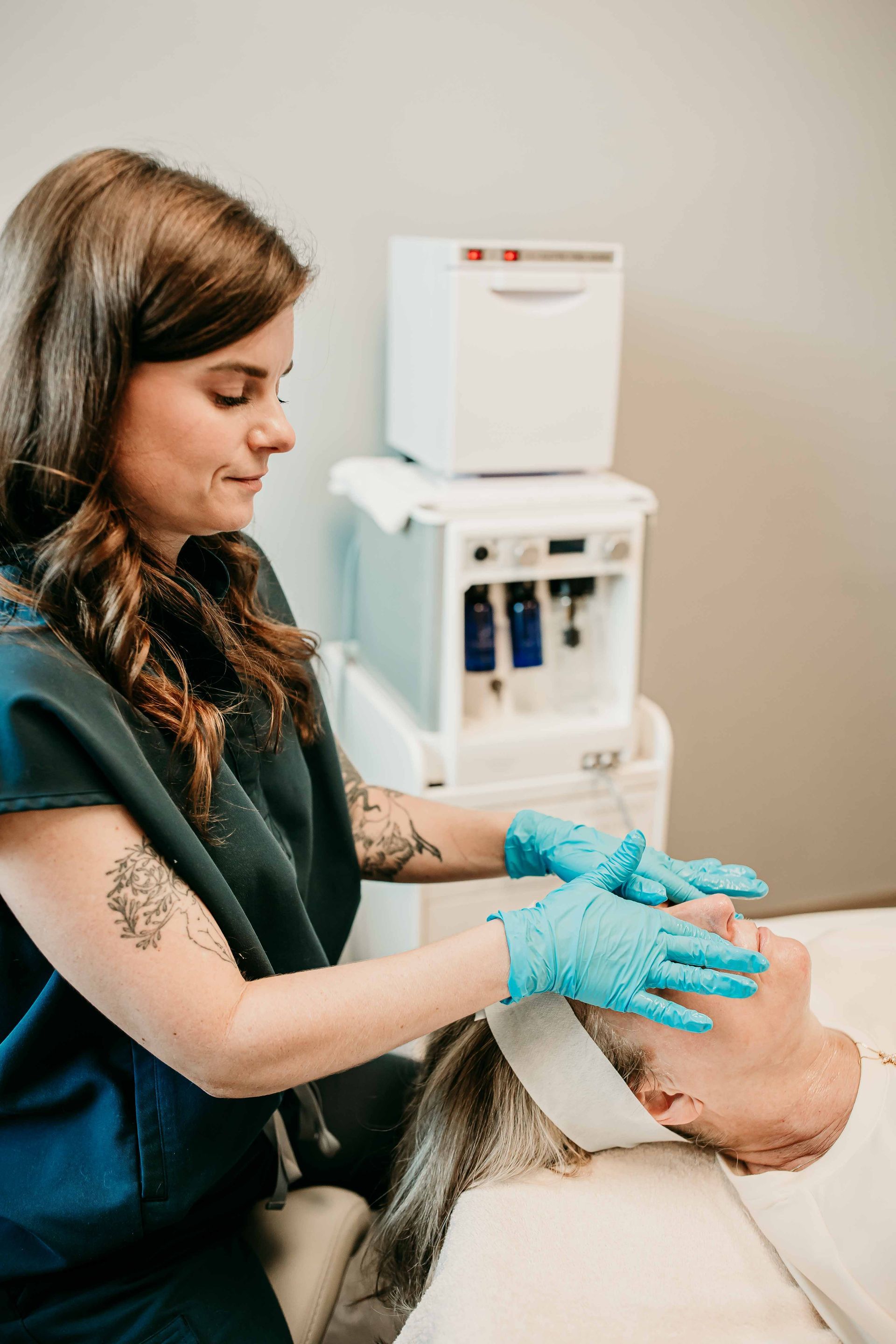 Esthetician in blue gloves performing facial treatment on client, beside skincare equipment.