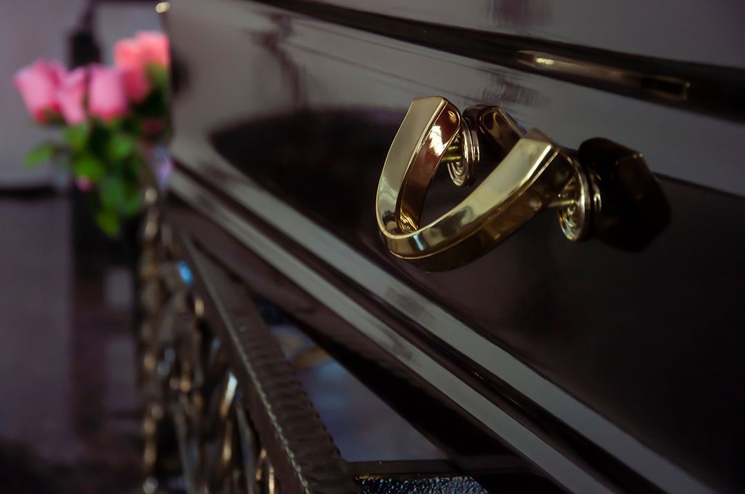 A polished dark brown casket with gold hardware in a room with a blurred bouquet of pink roses in the background.