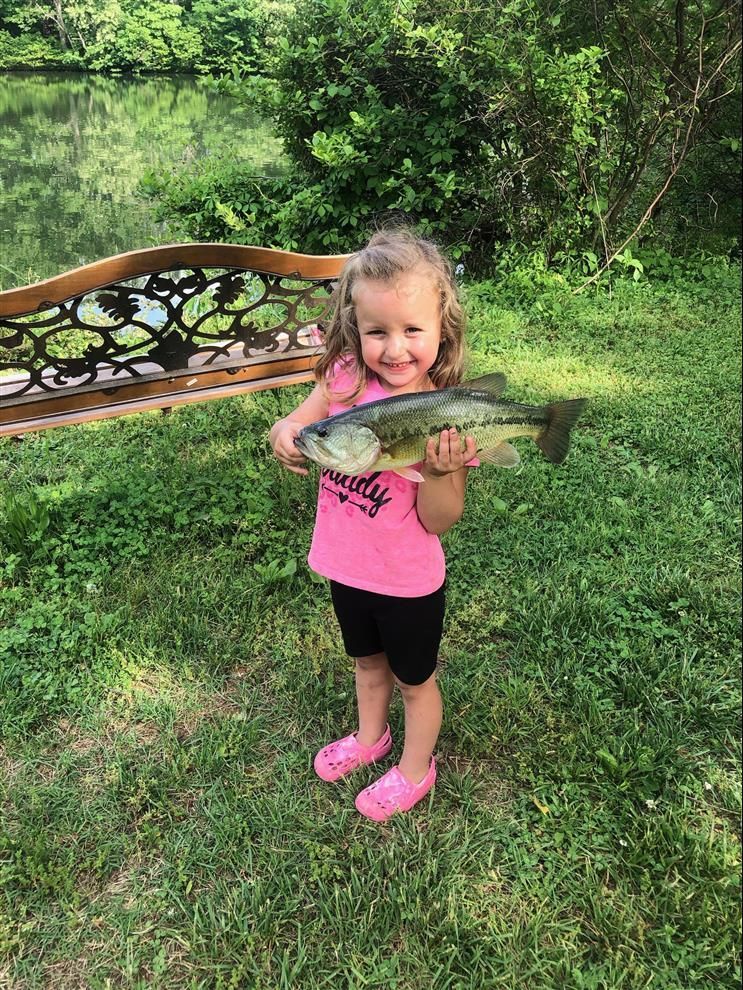 Young girl in pink shirt and crocs, holding a large fish, smiling, by a pond.