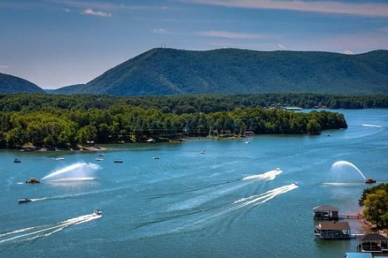 Boats on a lake with water fountains, surrounded by green trees and mountains under a blue sky.