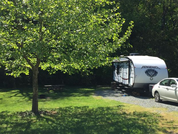 RV and car parked at a campsite, shaded by a tree. Green grass, gravel, and trees are visible.