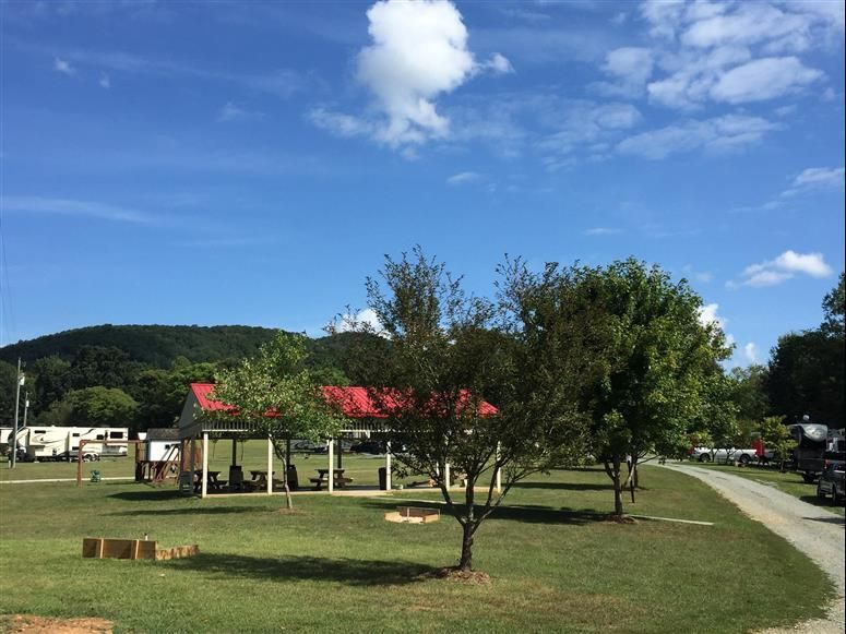 Campground scene with picnic shelter, RVs, trees, green grass, and a mountain backdrop under a blue sky with clouds.