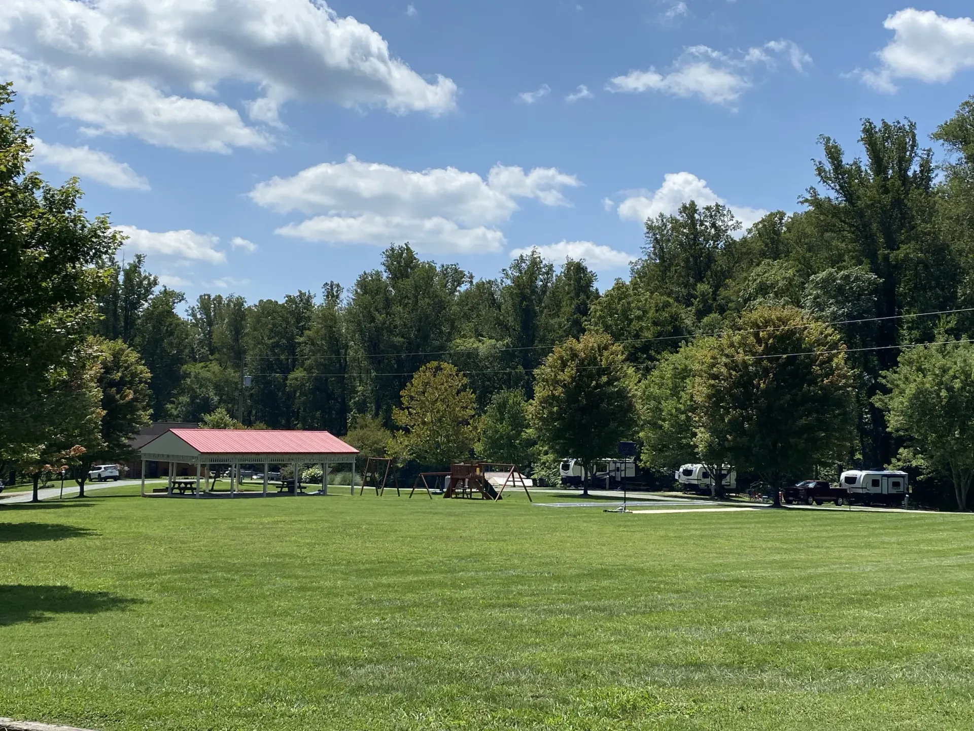 Lush green park with trees, picnic shelter, RVs, and sunny blue sky with clouds.