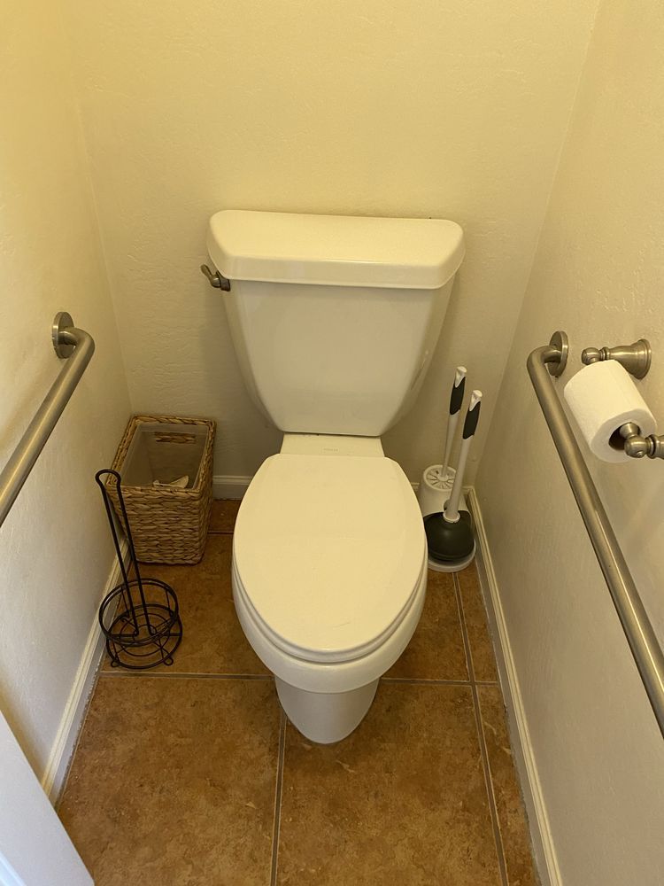 Toilet in a small bathroom with grab bars, toilet paper holder, and plunger.