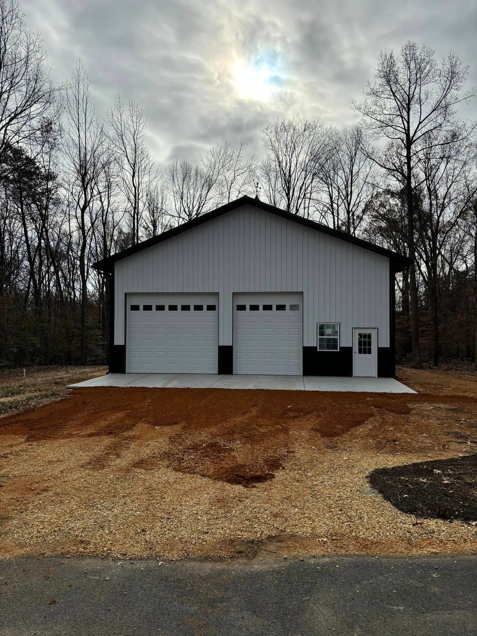 A white pole barn garage with two garage doors is sitting in the middle of a dirt field.
