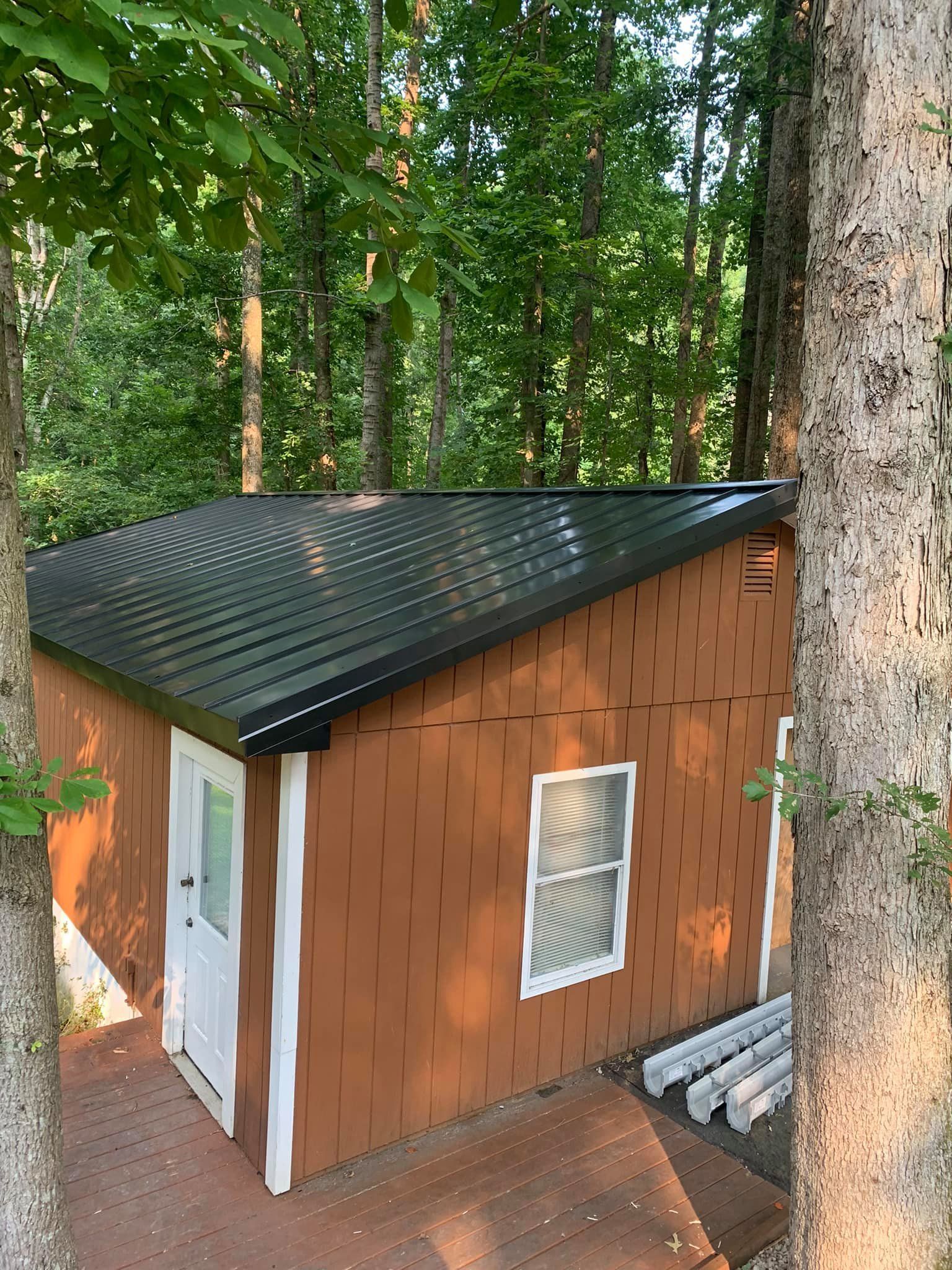 A small brown house with a black metal roof is surrounded by trees.