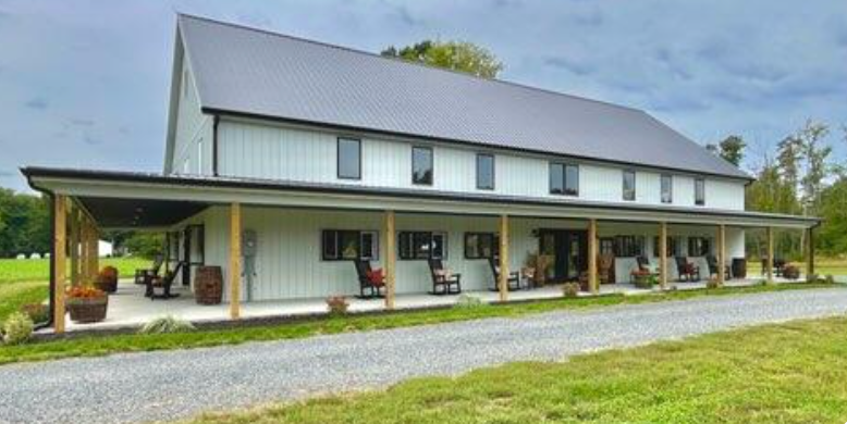 White barn-like building with a long porch, rocking chairs, and a gravel driveway.