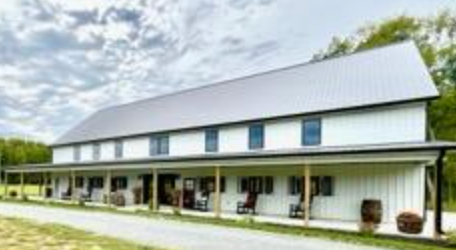 White barn with a metal roof and a long porch. Wooden barrels flank the porch. Cloudy sky background.