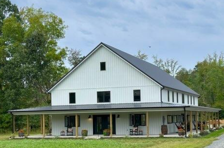 White two-story barn with a black roof and wraparound porch, set in a grassy area with trees.