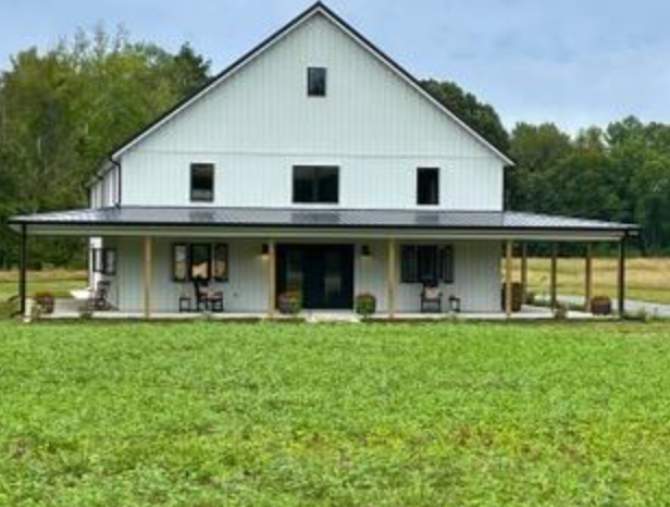 White farmhouse with black roof and porch, sitting on a green lawn.