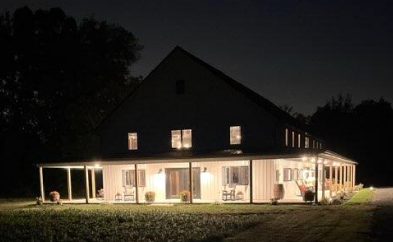 Two-story white house (metal pole barn)  with a wraparound porch at night, lit by porch lights.