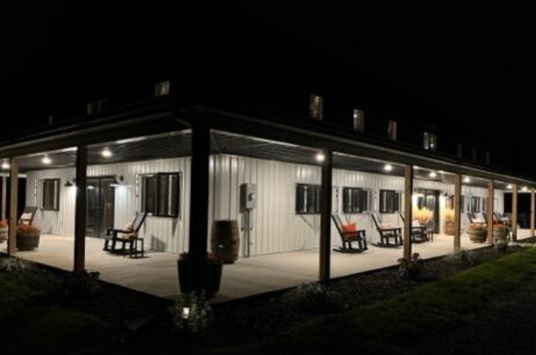 Lit-up barn with a wraparound porch at night. White exterior, rocking chairs, barrels, and dark windows.