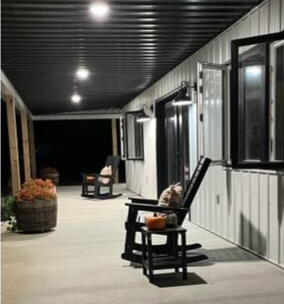 Black rocking chairs on a porch decorated for fall with pumpkins, barrels, and a dark corrugated metal ceiling.