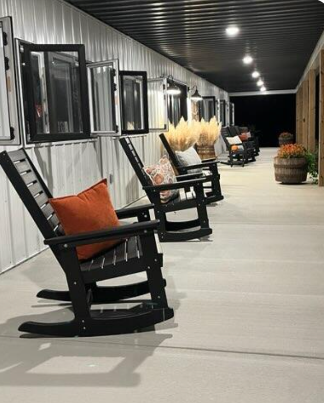 Row of black rocking chairs on a pole barn with pillows, wheat stalks, and a barrel; windows along the wall.