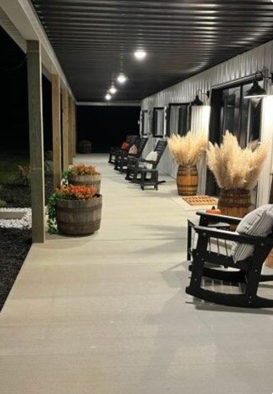A pole barn porch with rocking chairs, barrels, and pampas grass. Evening setting with overhead lights and dark surroundings.
