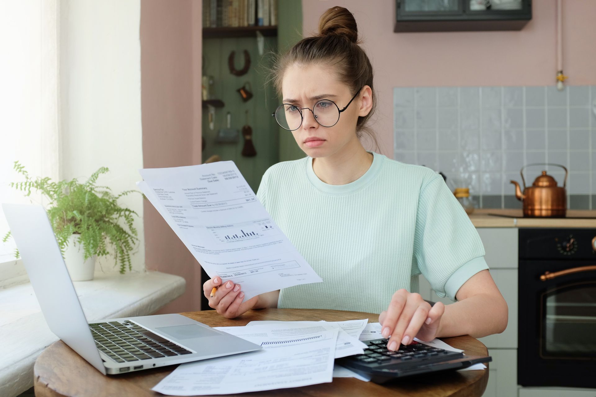 Woman with glasses using a calculator and looking at papers, likely bills, in a kitchen setting.