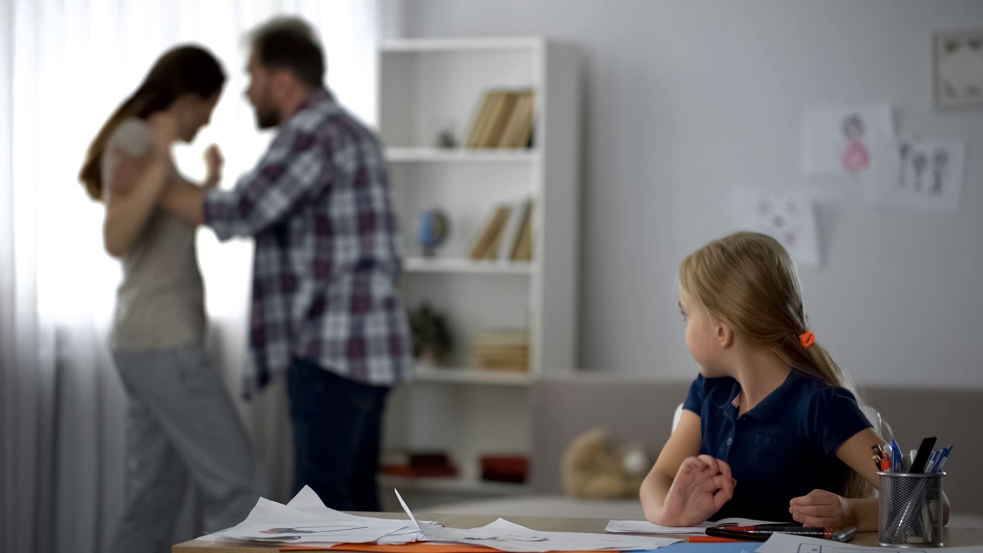A child watches as blurred parents argue in the background, papers on the table.