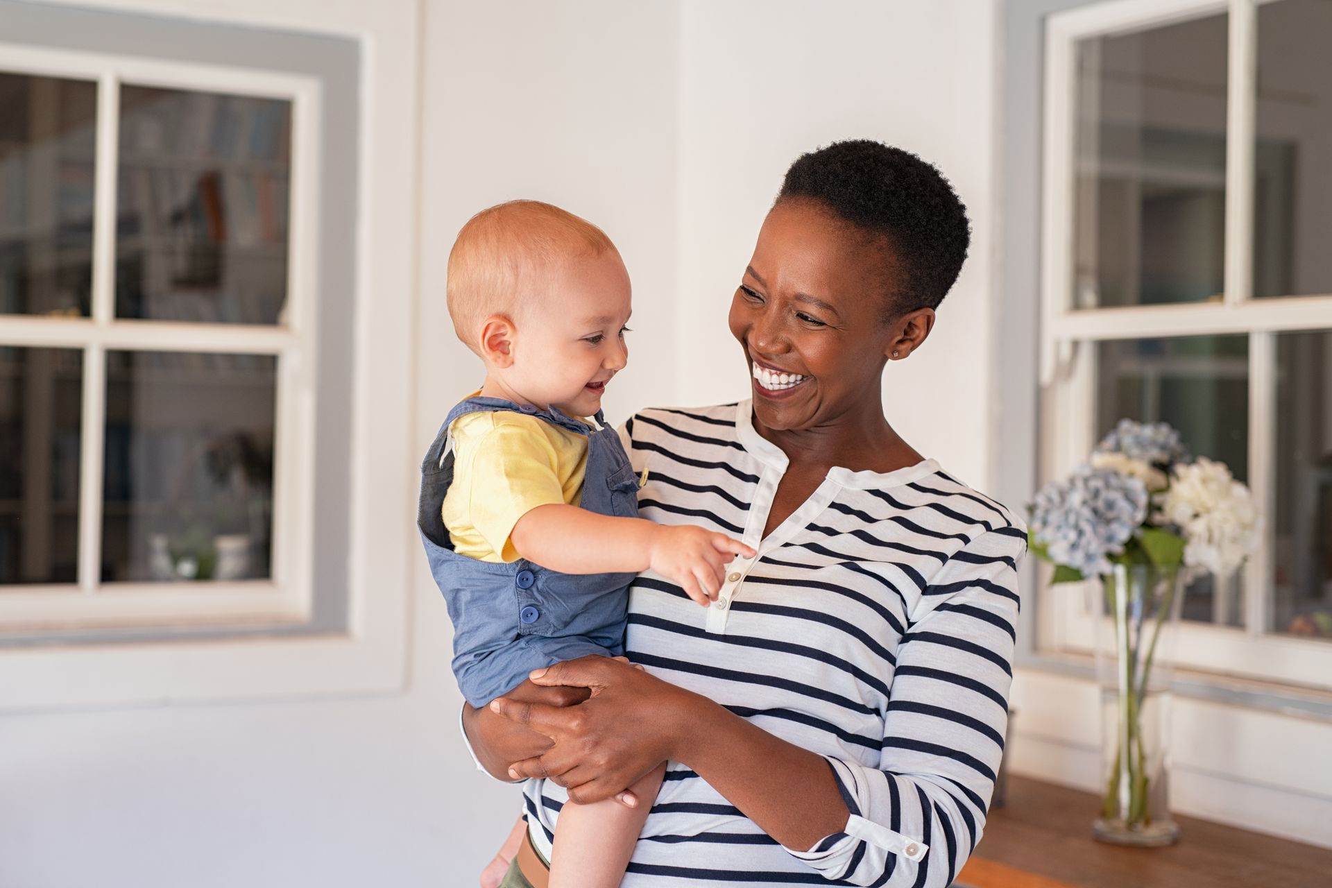 Woman holding and smiling at a baby in a room with windows and flowers.