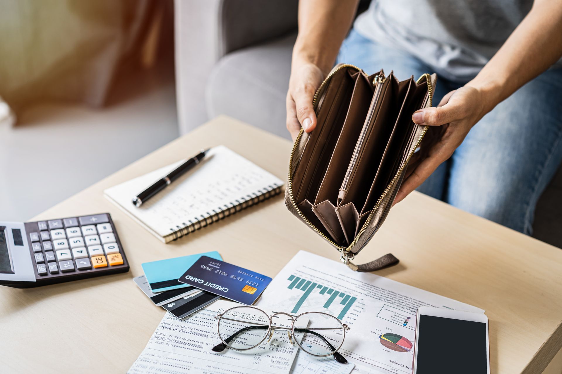 Person holding an empty wallet, surrounded by bills, credit cards, calculator, and notebook on a table.