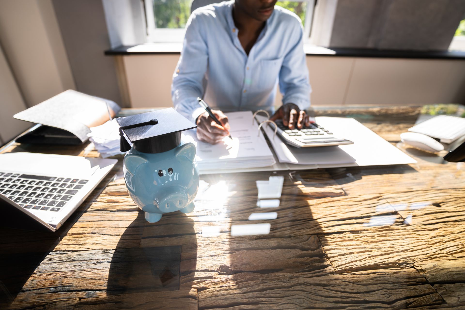 Person calculating finances at desk, piggy bank with graduation cap in front.