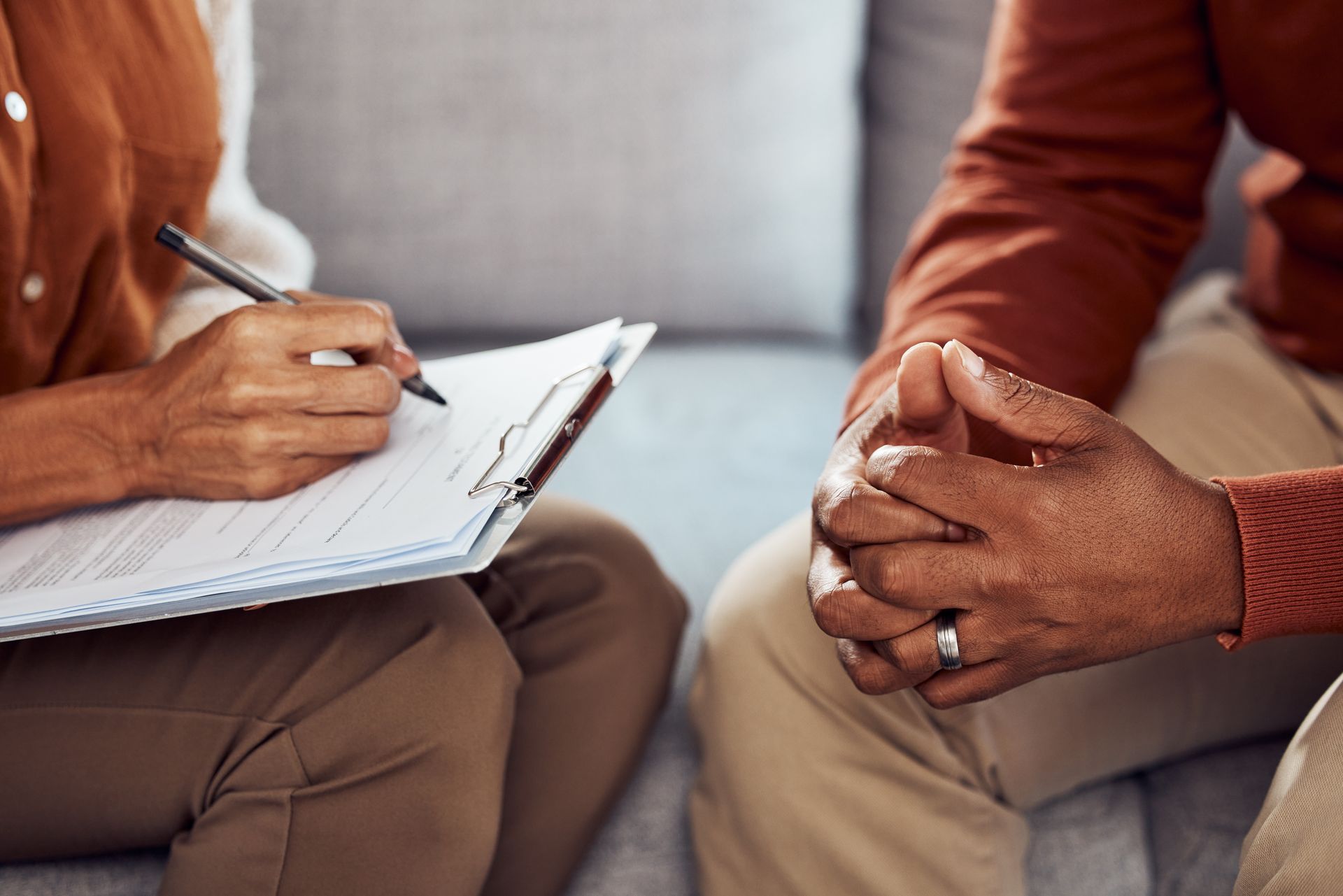 Therapist writing on a clipboard while the patient clasps hands, sitting on a couch.