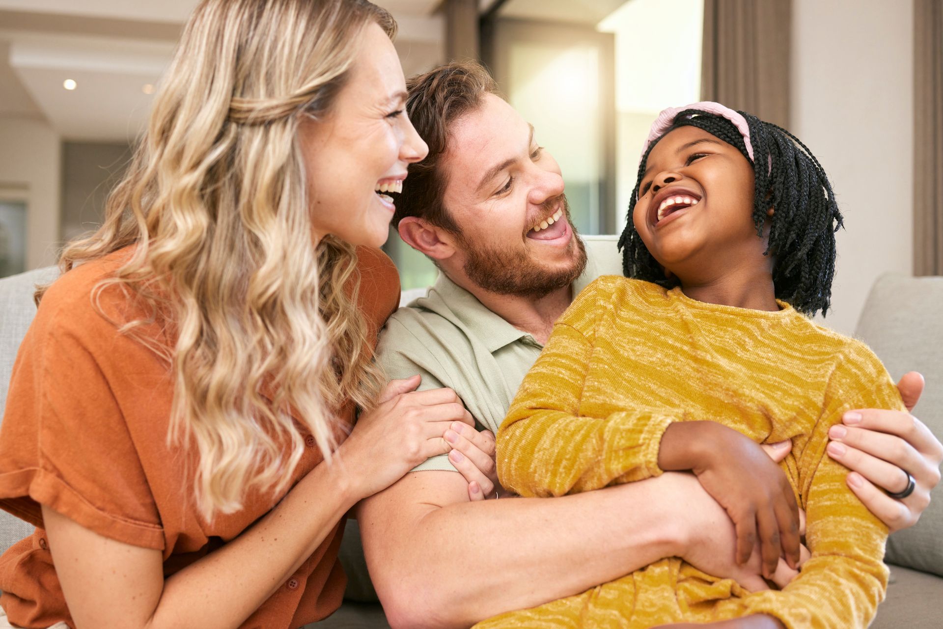 Family laughing on a couch. A woman, man, and child embrace. Sunlight streams through window.