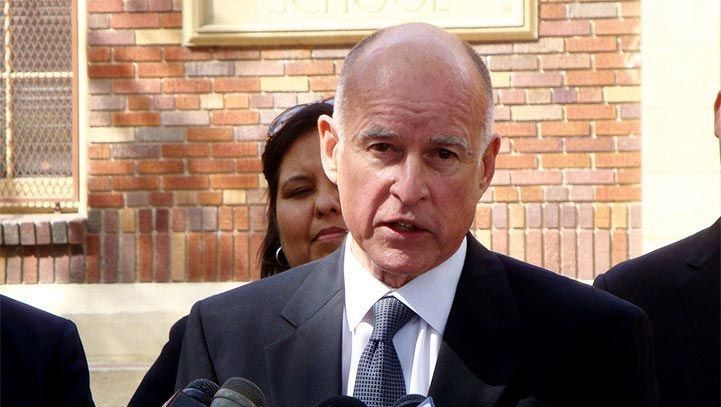 Man in dark suit speaking into microphones, brick building in background.